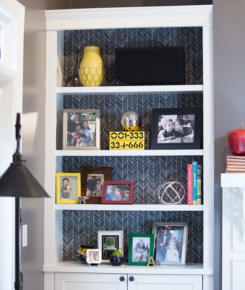 A white bookshelf with gray and white peel and stick wallpaper used at the back of it, stands against a gray wall. Family photos, a speaker, books and a vase are on the shelves. 