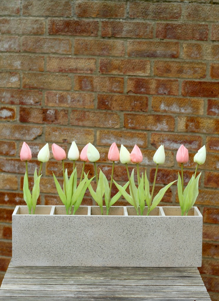 Pink and white tulips extend out of a long gray planter box from each of the five individual boxes within it. The planet is on a wooden table in front of a red brick wall. 