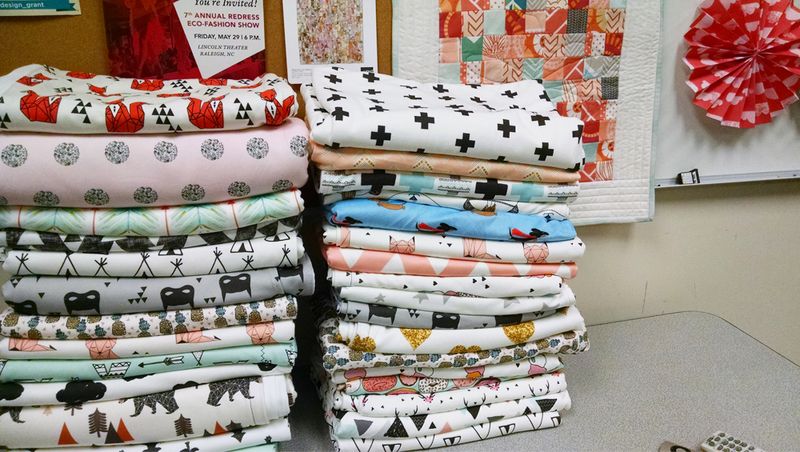 Two stacks of over a dozen blankets each sit on a table ready to be donated to charity. The patterns mostly have a white background, many feature small animals or small black geometric designs. 