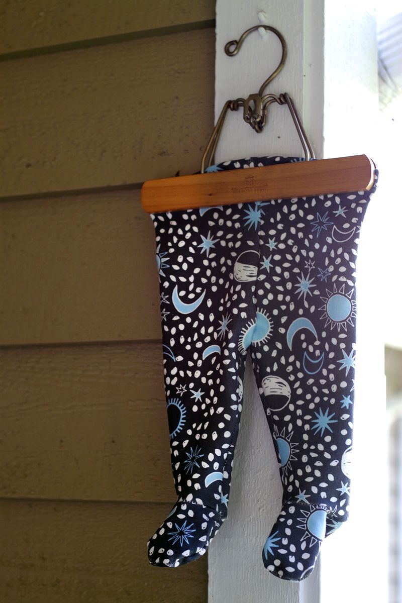 The footie leggings with a white-and-light blue sun, moon and star design on a navy background are hung up with a wooden hanger against the wooden side of a building. 