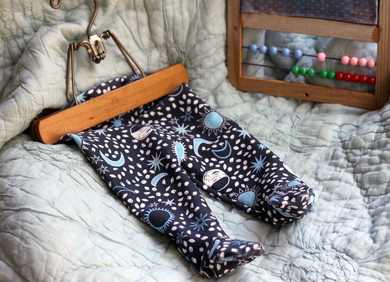 An image of finished footie leggings with a white-and-light blue sun, moon and stars design on a navy background. The leggings are on a wooden hanger and lay on a gray blanket next to a child's toy with pastel beads