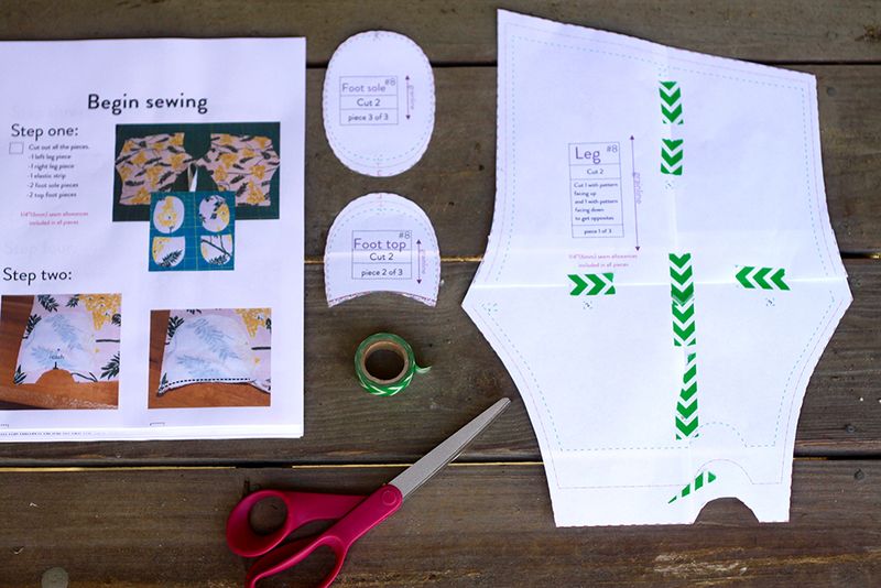 The footie leggings pattern is cut out with a part for the legs, the foot top and the foot sole. Red-handled scissors and the pattern's instructions lay beside the pattern pieces. 