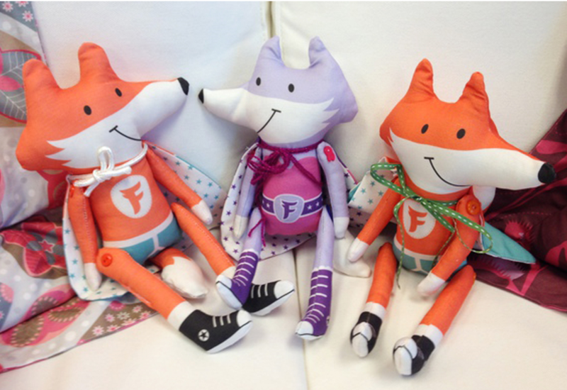 Three handmade foxes sit on a white sofa. Two foxes are orange and white and wear black high-top shoes and one fox is lavender and white and wears purple high-top shoes. They all have a "F" on their chest and are wearing superhero capes. 