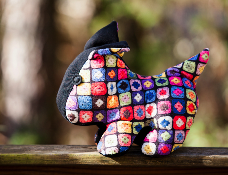 A small plushie of a dog made from fabric with granny squares sits on a wooden rail. 