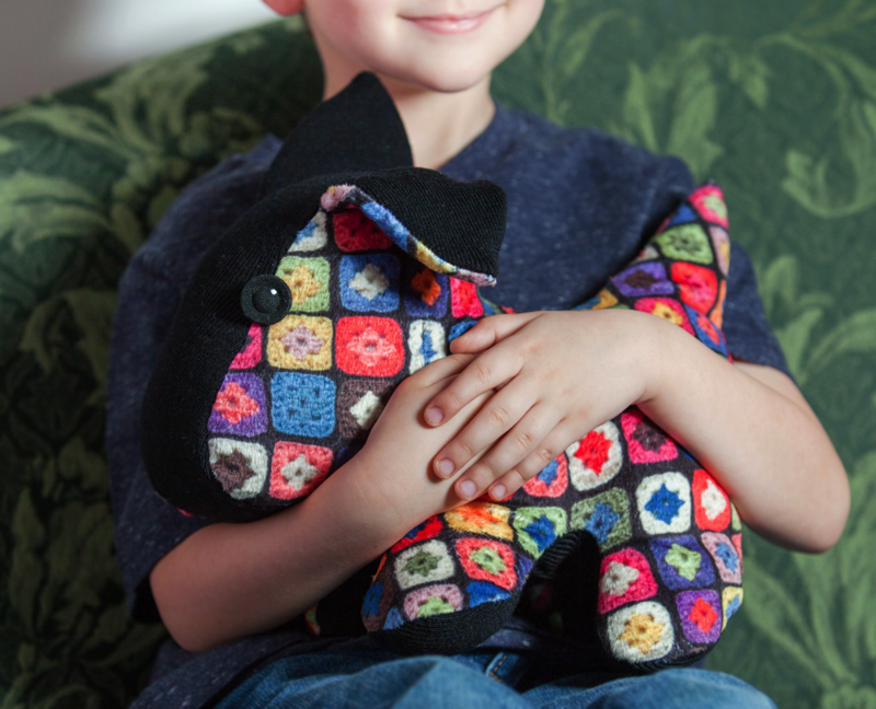 A child is holding the finished Granny Square Dog Plushie and sitting on a green couch. A smile is on the child's face. 