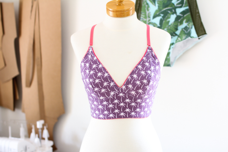 A dark purple bralette with small white palm trees and hot pink straps is on a white sewing form