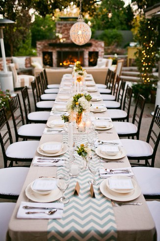 A long table is covered in a beige tablecloth with a dusty blue and white table runner. Off white plates with bowls on top of them and silver cutlery on either side on white napkins run in place settings down both sides of the table. A fireplace is at the far end of the table. A black chair with a white cushion is by each place setting. 