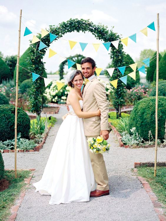 A couple stands underneath turquoise, yellow and white bunting on a gravel path in with a garden featuring shrubs on flowers on either side 