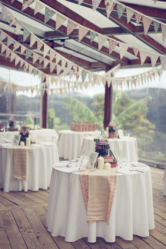 Small tables with cutlery, beige table runners, candles, a birdcage fill of flowers and a white tablecloth sit underneath rows of gray, white and brown bunting hung from the ceiling 
