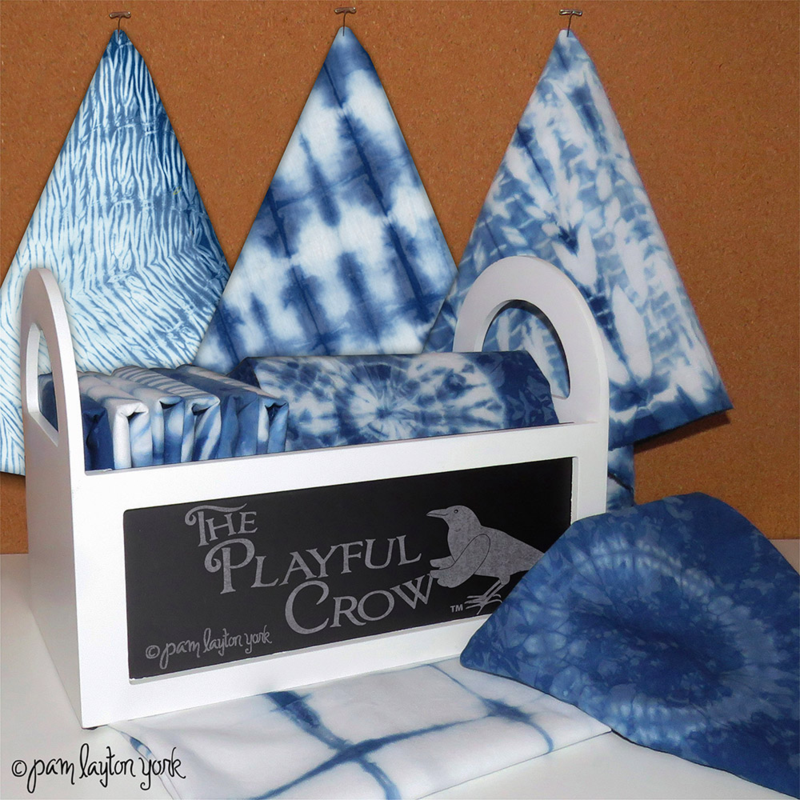 Three blue-and-white shibori napkins are pinned to a corkboard with t-pins. The are behind a white box with rounded handles full of folded shibori napkins. Additional shibori napkins lie beside the box. The front of the box has been painted black and has a white crow design on it and large white text that says "The Playful Crow." @pam layton york is written to the bottom lefthand side.