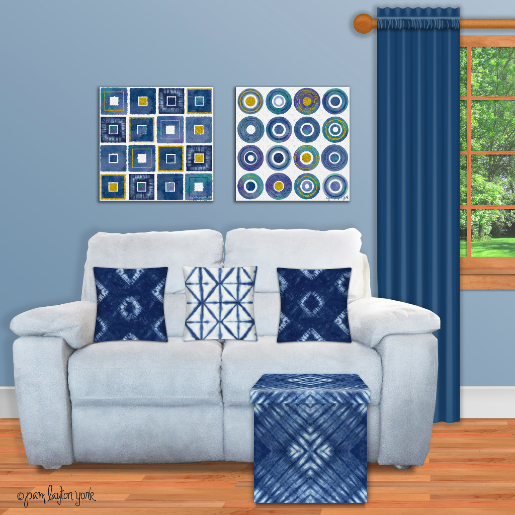 A photo of a render of Pam's designs, i.e., what they would look like in someone's home. There is a white couch with three shibori blue-and-white pillows on it and a blue-and-white shibori box in front of the sofa. On the wall behind the sofa are two pieces of art. The one on the left has 12 small blue-and-white shibori pieces in it, arranged in 4 rows of 4 fabric pieces. The piece of art on the left is similar, except that the squares of shibori fabric are circles. There is a dark blue curtain panel hanging to one side of the window to the right of the couch.
