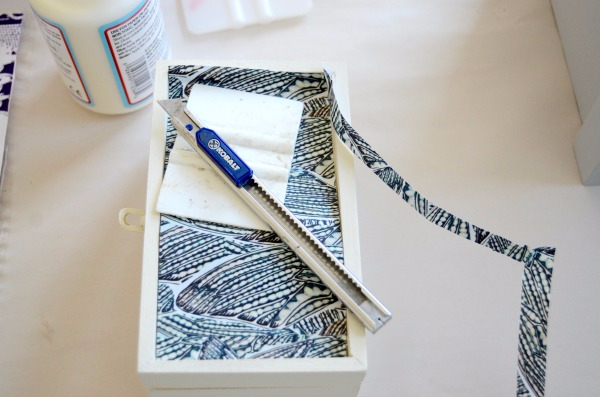 A piece of wallpaper with blue-and-white butterfly wings on a white background is being cut to the same size of a white jewelry box. 