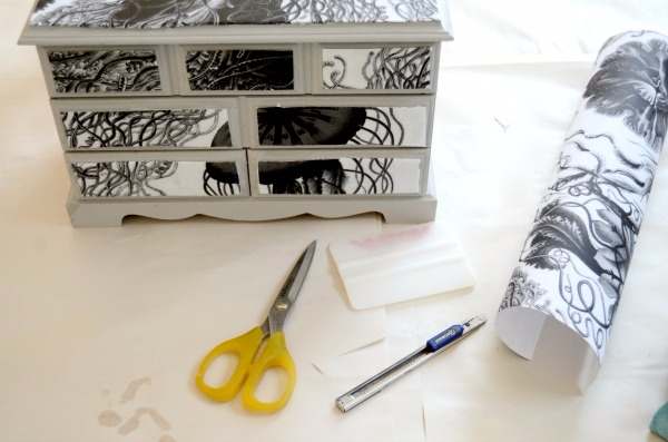 A small gray jewelry box has the top and small front drawers covered in black-and-white jellyfish wallpaper. Extra jellyfish wallpaper, scissors and an x-acto knife sit around the jewelry box. 