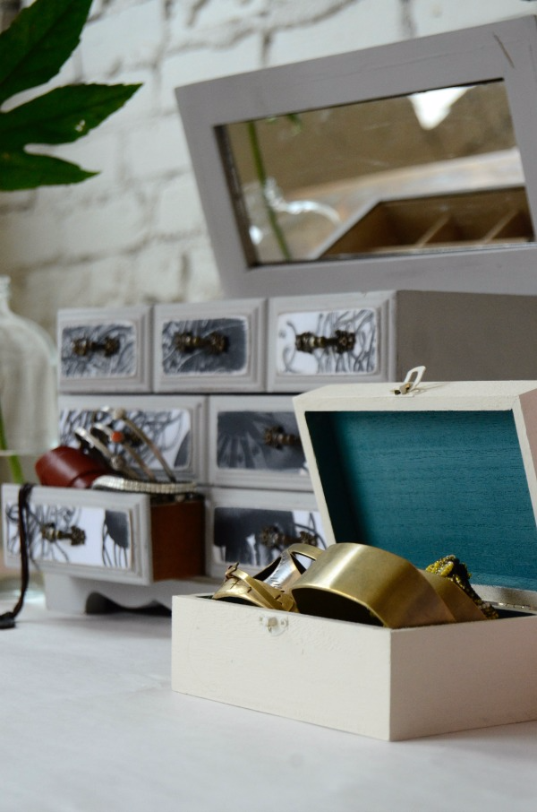 A gray jewelry box with black-and-white jellyfish wallpaper on the front drawers sits next to a smaller white jewelry box with a teal interior and gold bracelets spilling out of it. 