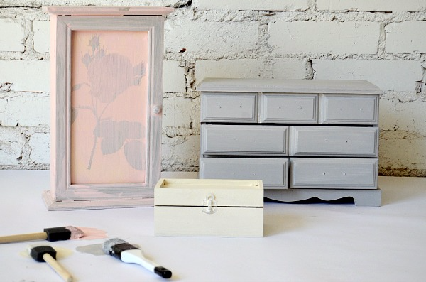 Three small jewelry boxes are on a white table, one painted light pink, one painted white, one painted gray. Three small paint sponges, one with pink paint on it, one with gray paint, one with white paint lay in front of the jewelry boxes. 