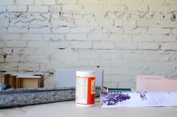 A roll of gray-and-white wallpaper and roll of mostly white wallpaper with a bright purple design at the bottom lay on a white table next to a bottle of Mod Podge. 