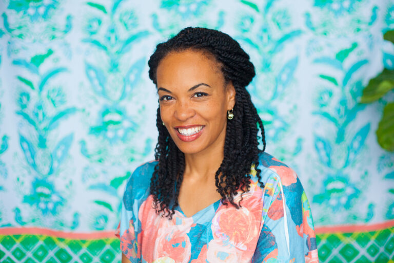 woman with black braids and a blue and pink top sitting in front of a blue floral background