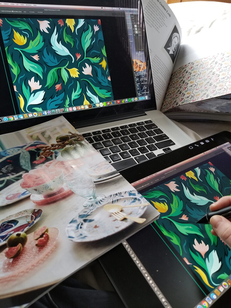 Claire drawing on a tablet that is showing up on a computer screen in front of the tablet. A magazine spread featuring a table with plates set around it and figs on the table too. The design Claire is drawing features green, pink, yellow and red leaves floating through a navy background. 