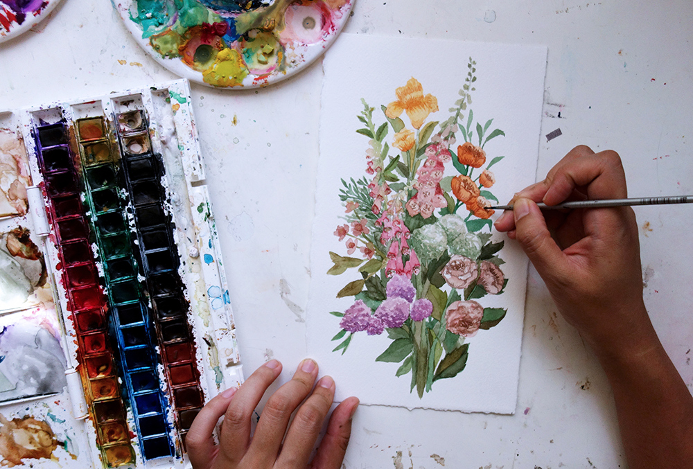 Cecilia is putting the finishing touches on a painting full of brightly color flowers collected in a bunch. A tray of 45 paints in small open sections lies to her left. 

