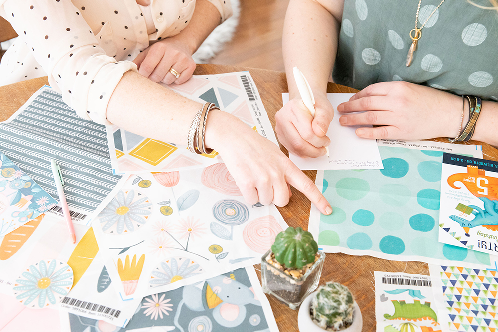 Jenica & Danielle's hands are in this photo with several of their designs in front of them on a table with two small succulents. The designs have been printed on paper and the pieces of paper are spread over the table. There are blue dots in varying shades on a light blue background, gray stripes, a piece of paper just showing a gray background with part of the face of a gray mouse, a piece of paper with pink and gray flowers on a white background. 

