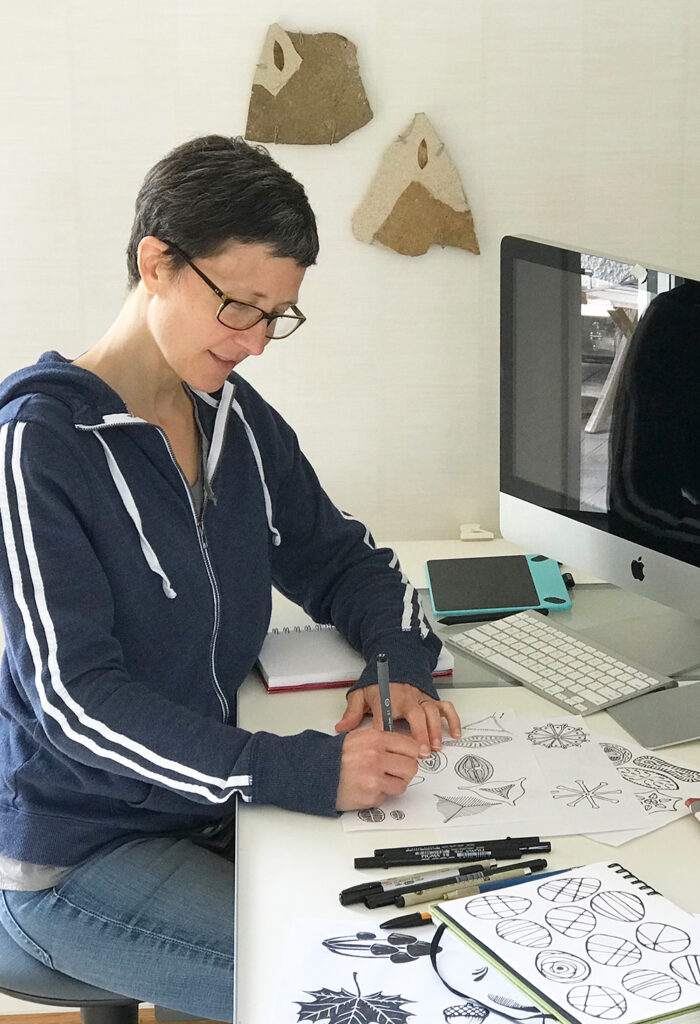 Allison is drawing small geometric designs on a white piece of paper with a black pen in front a computer monitor on a white desk. 
