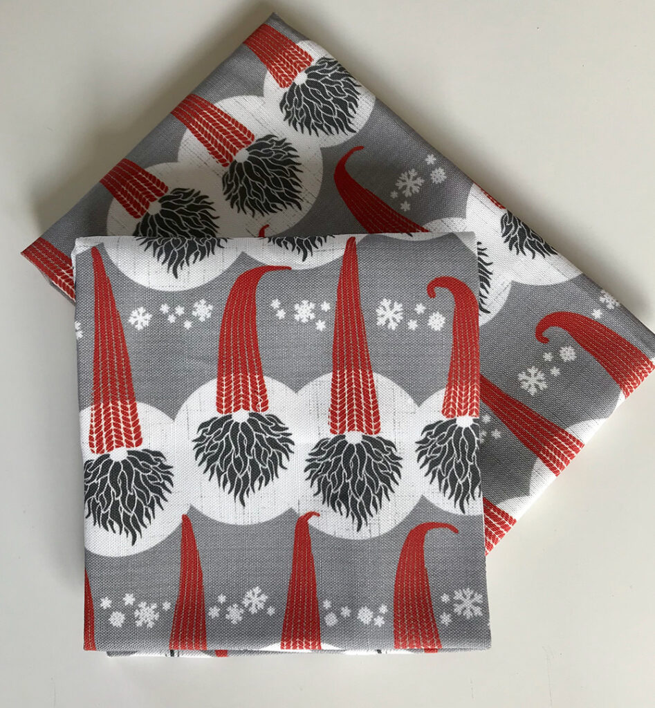 Allison's design Swedish Tomte on dinner napkins. The noses and black bushy beards of gnomes are shown popping out under tall thin knitted red hats on a gray background. White circles are shown over and under their faces and on the parts of the hats just above their noses. Clusters of small white snowflakes are between the tops of the hats.
