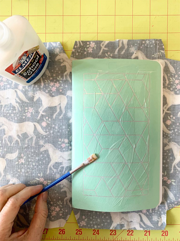 A paintbrush is over top of a journal that is covered in school glue. A piece of fabric is beneath the book. A yellow cutting mat is in the background.