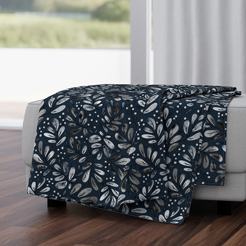 Daria's design Indigo Floral Snowberries, which has a navy background and small white flowers dotted all over, as seen as a blanket thrown over a gray couch in front of a large window. 