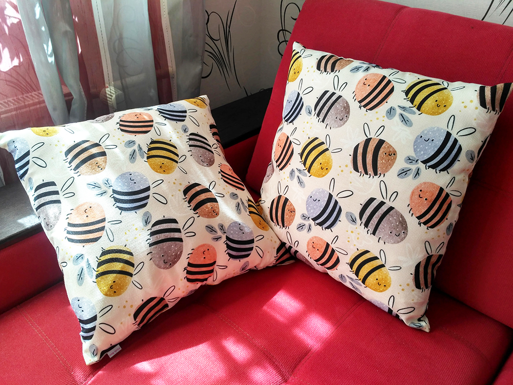 Daria's design Watercolor Bees Pattern on two pillows on a red chair. The design has a cream background and round bumblebees that are blue, red, yellow and purple, all with three thick black stripes on their bodies, flying around it. 
