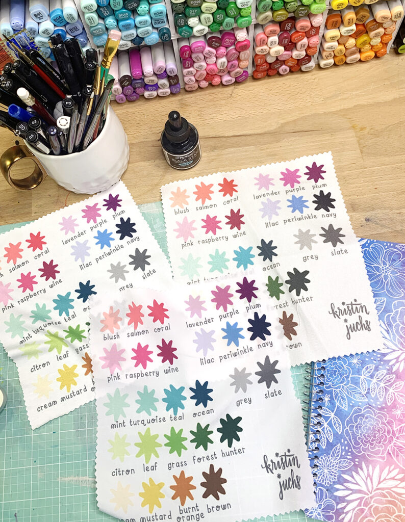 A photo of color palettes Kristin has made for the colors that are designs use. The color palettes are printed on pieces of fabric, which are laid out on a wooden desk. 