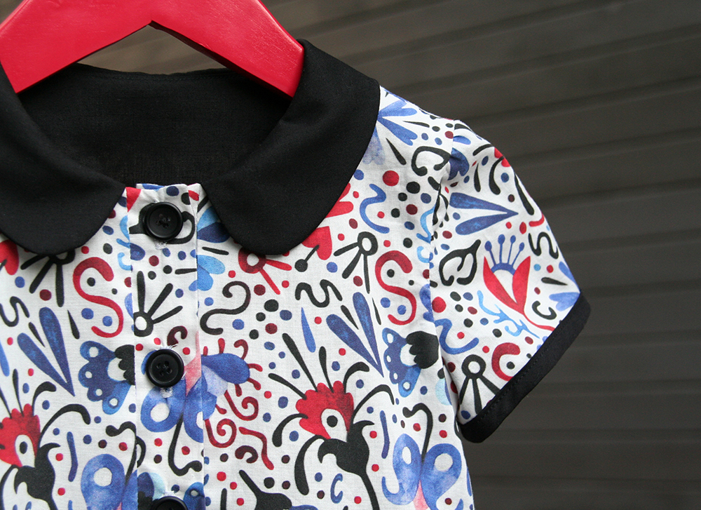 Marta's design Red and Blue Party as seen on a small shirt with a black collar, buttons and sleeve edge. The design has a white background and red and blue flowers on black stems. 

