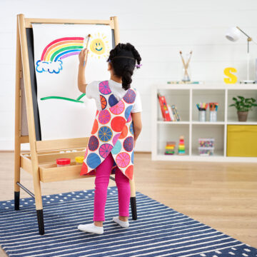 Girl painting a rainbow on an easel while wearing a handmade, colorful art smock