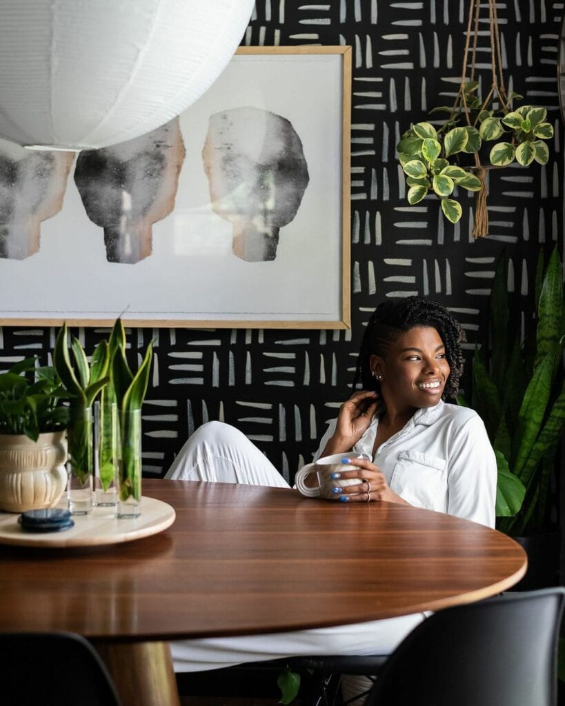 A women with a coffee cup sits at a dining room table in front of a wallpapered wall featuring a black and white pattern