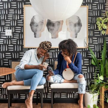 Two women sit on chairs and laugh at a cell phone in front of a black and white patterned wallpaper wall