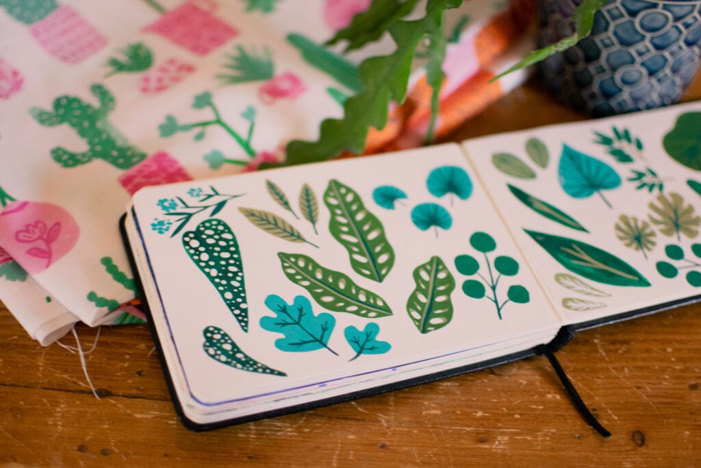 An opened sketchbook with drawings of leaves with a live plant and fabric with printed houseplants on it in the background
