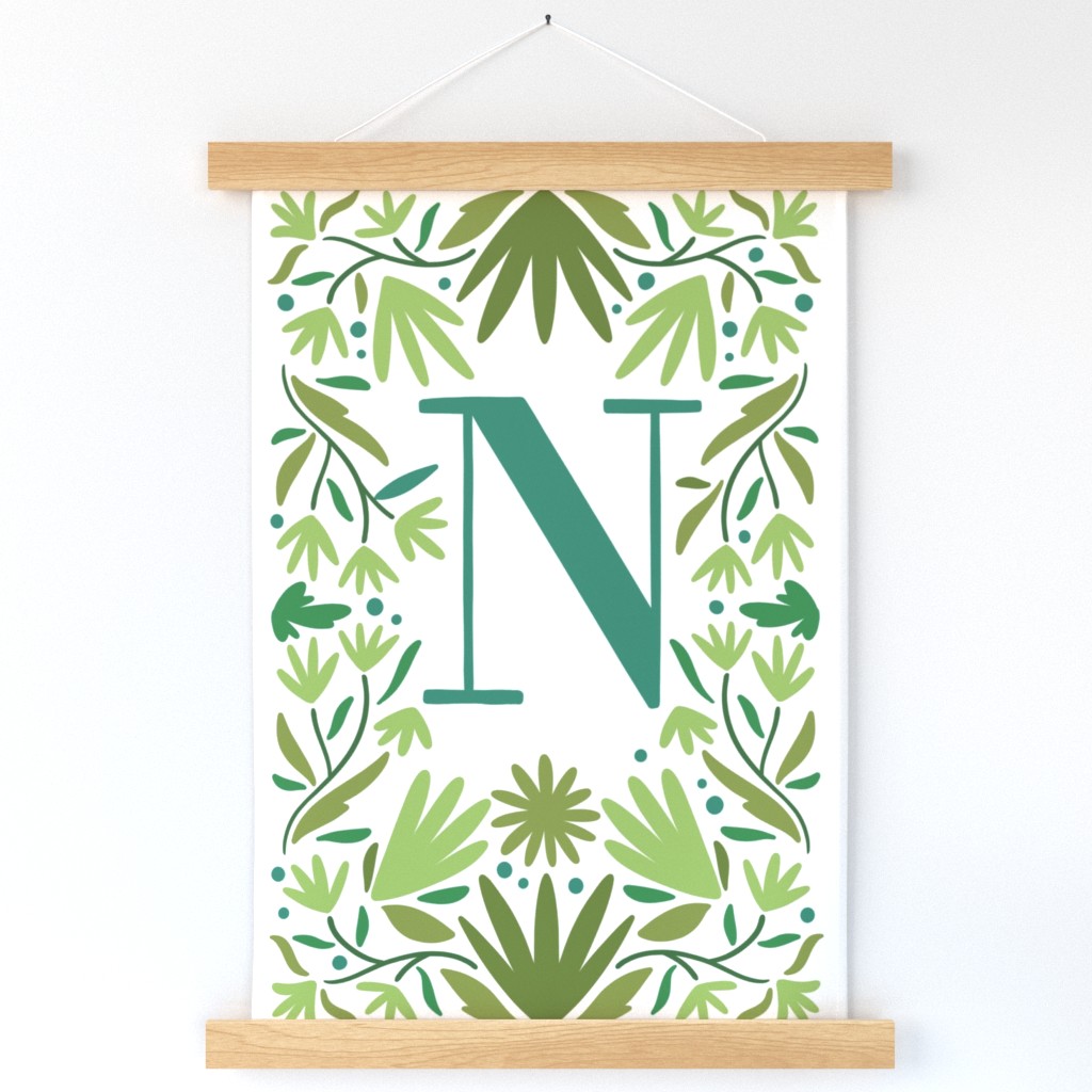 A wall hanging with a large green "N" surrounded by green flowers and branches hangs on a white wall.