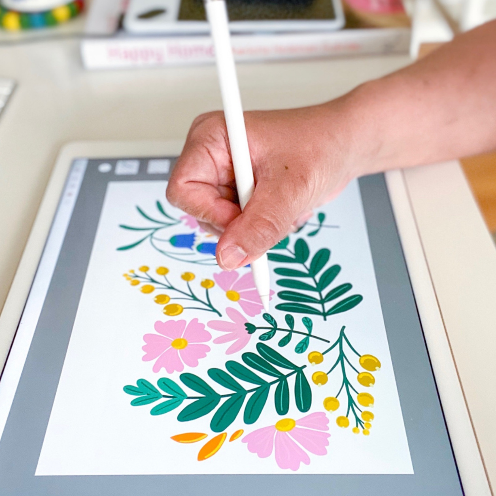 Close up of a hand drawing a design with pink flowers, gold berries and green leaves on a tablet with a digital pen