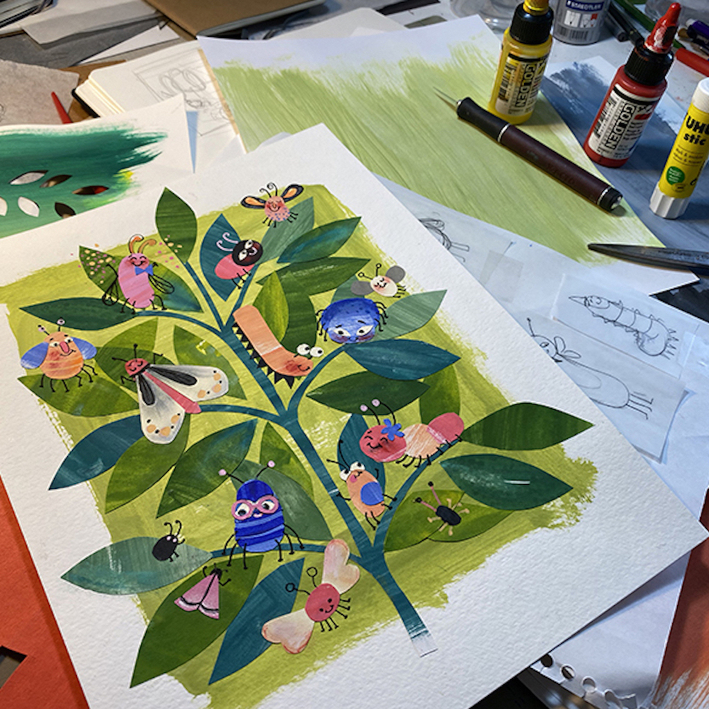 A drawing of a tree with green leaves filled with colorful bugs lies on a desk on top of colorful paper and next to a glue stick, pen and scissors.