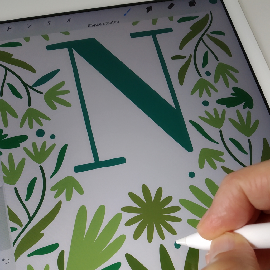 A close up of a hand holding a digital pen drawing green flowers on a design with a large dark green letter "N"