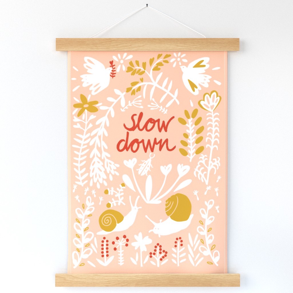 A pink wall hanging, with white snails and doves with gold accents surround the handwritten words "Slow down" in red on a pink background, hangs on a white wall