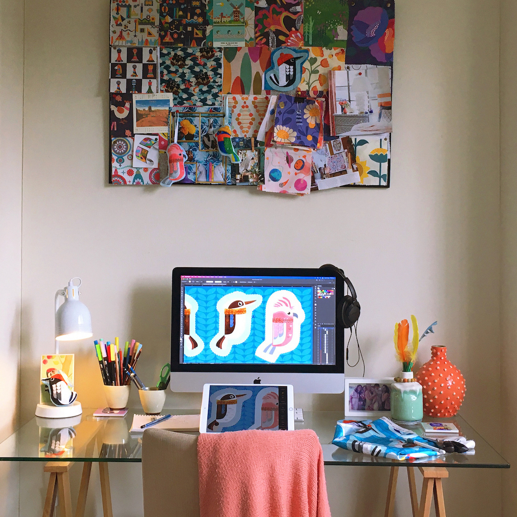 A laptop with a drawing of colorful birds on the desktop sits on a glass-topped desk below a cork board covered in bright geometric designs.