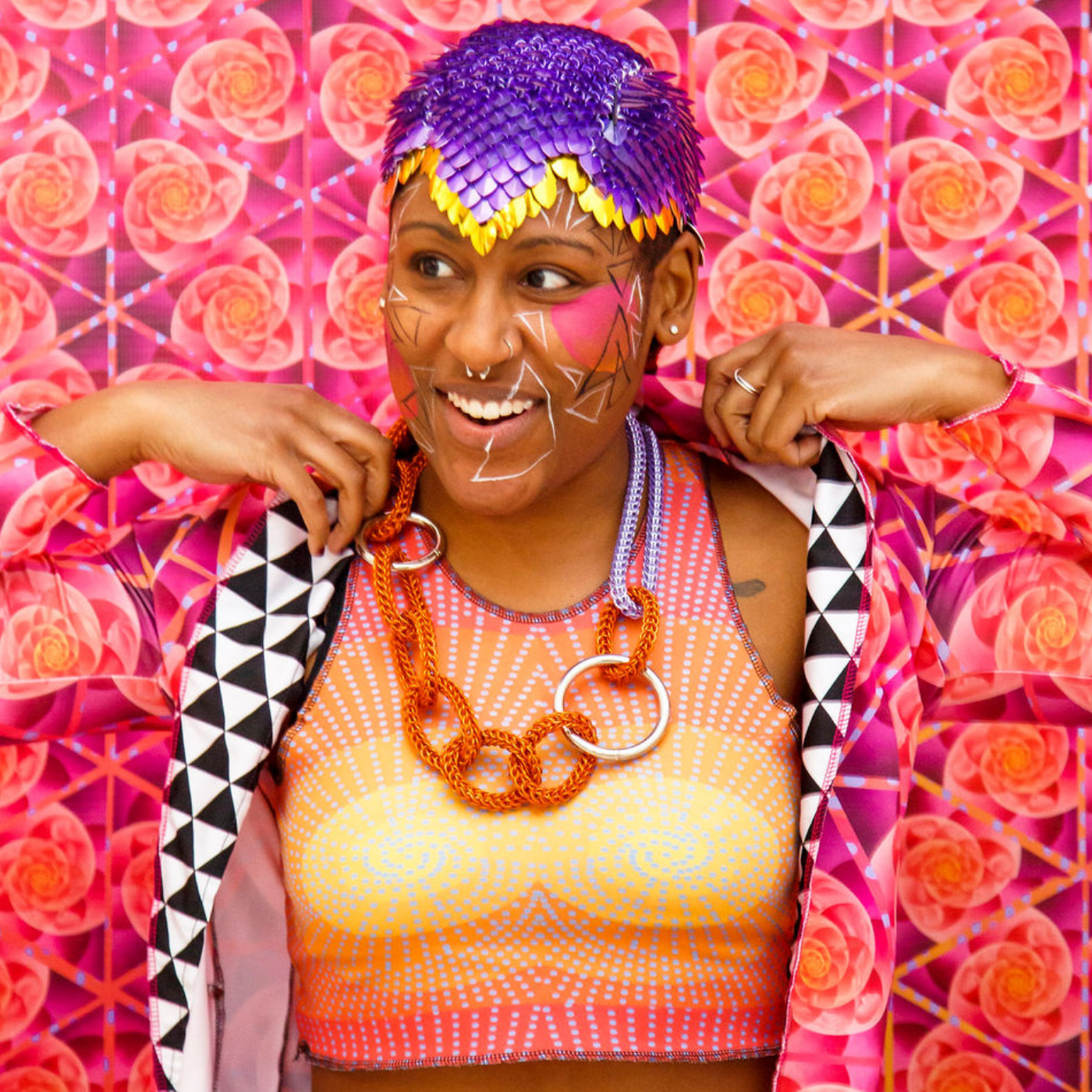 Ashley Olivia Morton stands in front of a bright orange and magenta wall with swirls, wearing a vest in the same pattern.