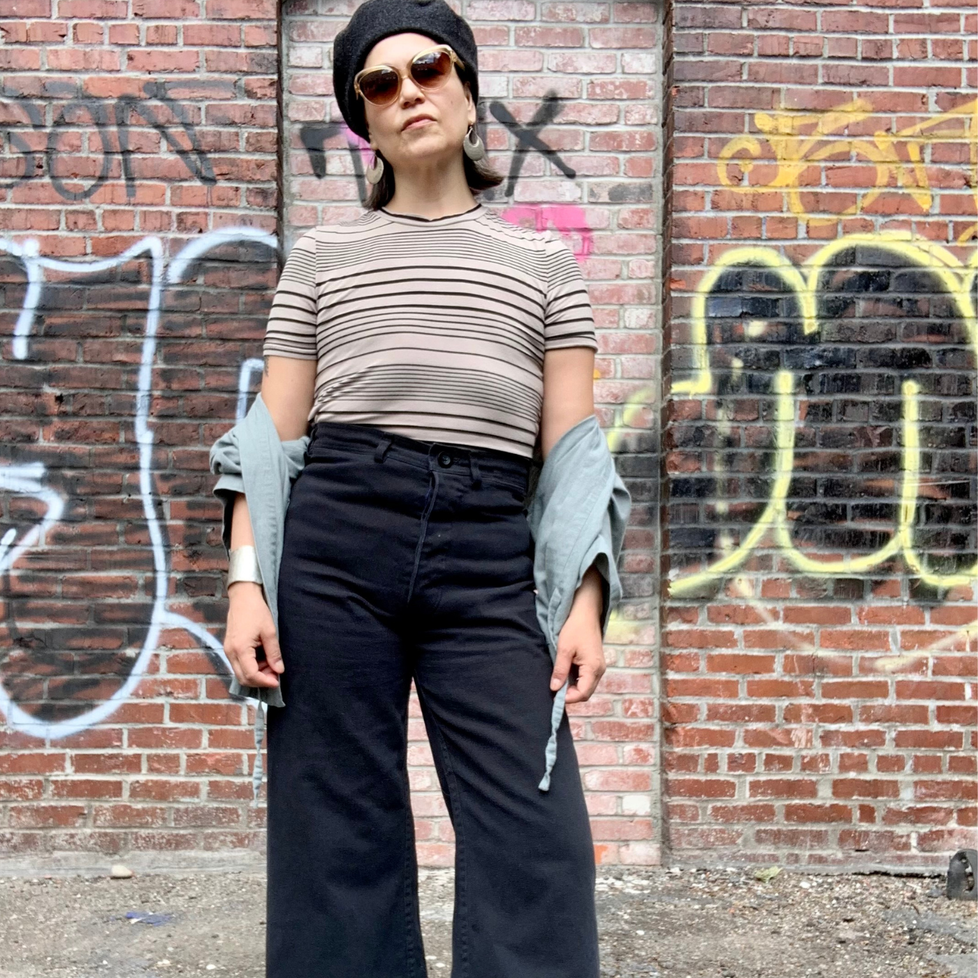 Denise Archer stands in front of a brick wall with graffiti wearing black trousers, a rose and black striped shirt, sunglasses and black hat