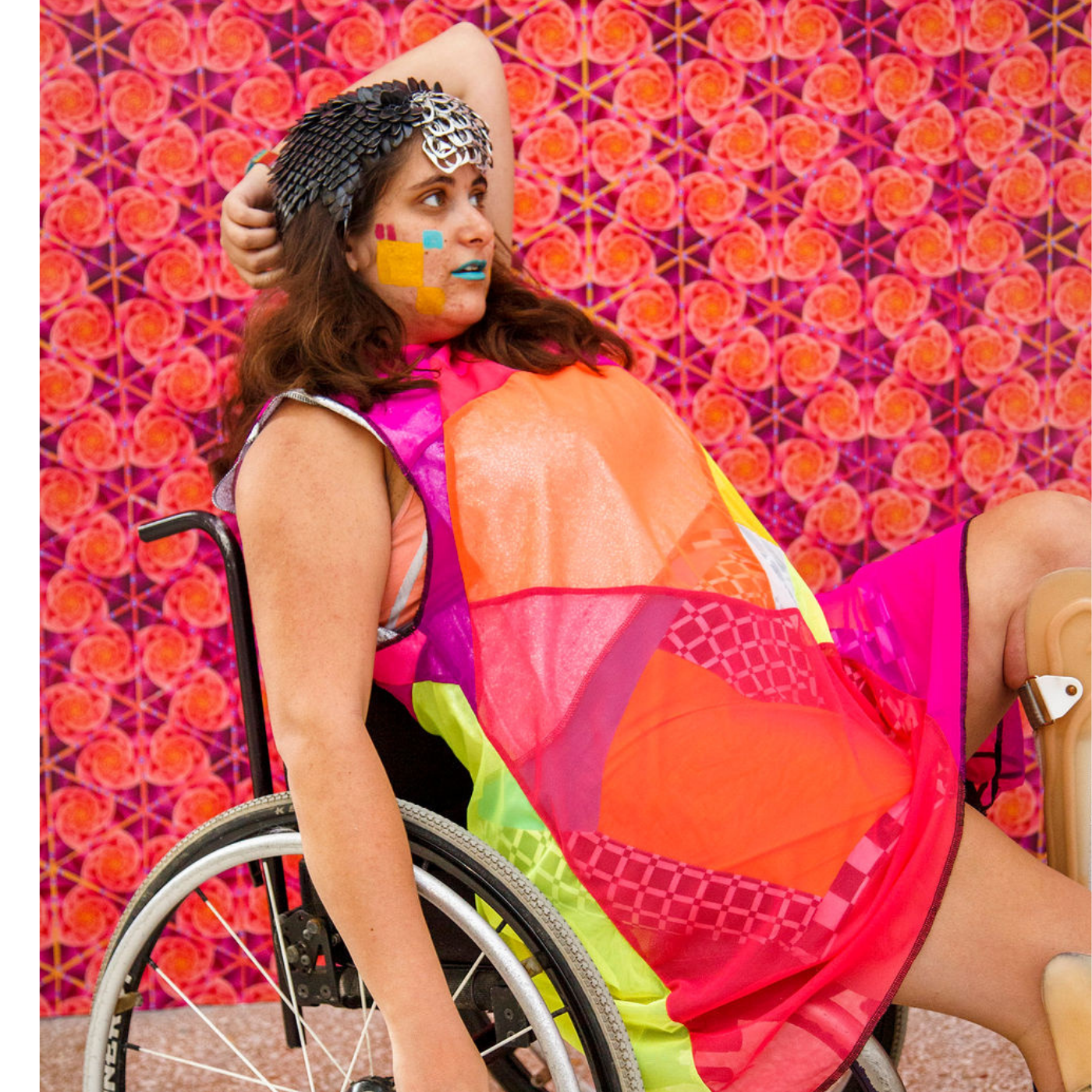 Emily Ash sits in a wheelchair in front of a bright orange and magenta wall with swirls, and leans back, left arm behind head and right arm pointing straight down.