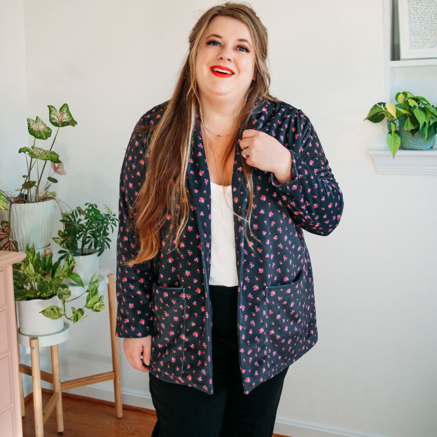Meg stands wearing black pants and a white top with a dark blue velvet blazer over top. The fabric of the blazer has small pink flowers dotted all over it.