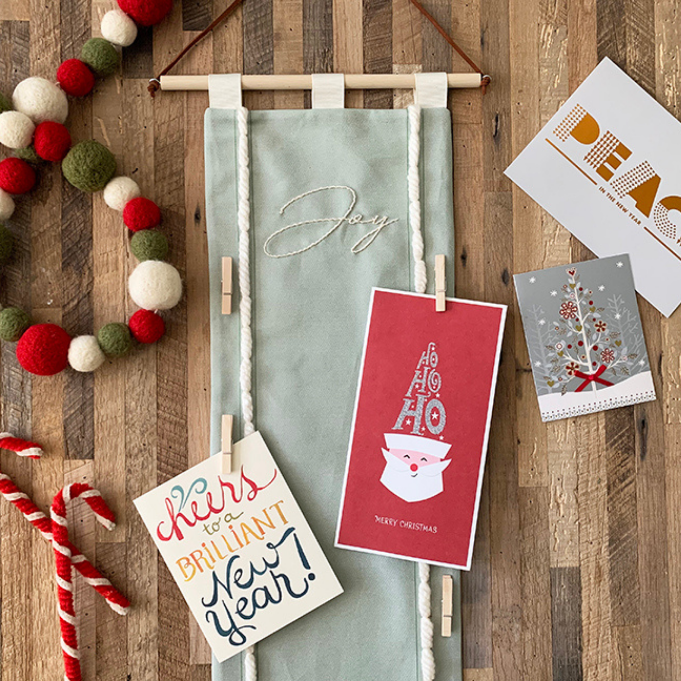 A fabric holiday card holder, which is sage green with the word Joy in white cursive font embroidered on it, has cheery holiday cards, one with the face of Santa Claus and another that says “Cheers for a Brilliant New Year!” in cursive red, yellow, green and blue, clipped to it with small wooden clothes pins. The card holder is encircled by various holiday things: red-and-white-fabric candy canes at the bottom left; red, cream and green felt garland at the top left; and a few holiday cards on the top right, one with a Christmas tree design and one with the word “PEACE” in red and gold all caps text. The card holder has a wooden dowel rod running at the top of it with brown twine coming up from either side, from which it will hang from the wall.