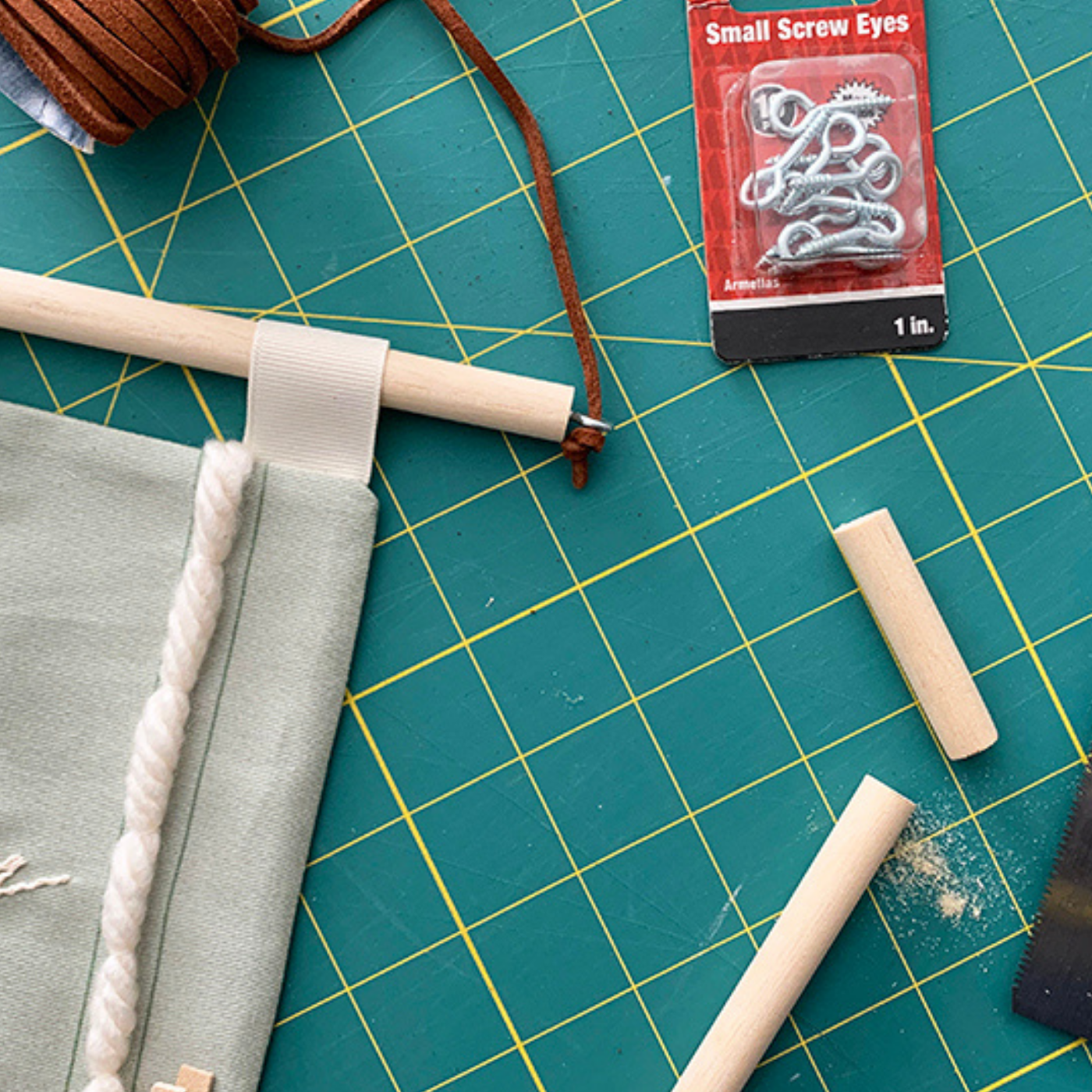 A close up of materials used to make a holiday card holder, some sawed-off bits of a small wooden dowel rod, a package of small white screw hooks, and a spool of brown twine, sit next to the holiday card holder itself, which is made of fabric and has a wooden dowel rod running across the top.