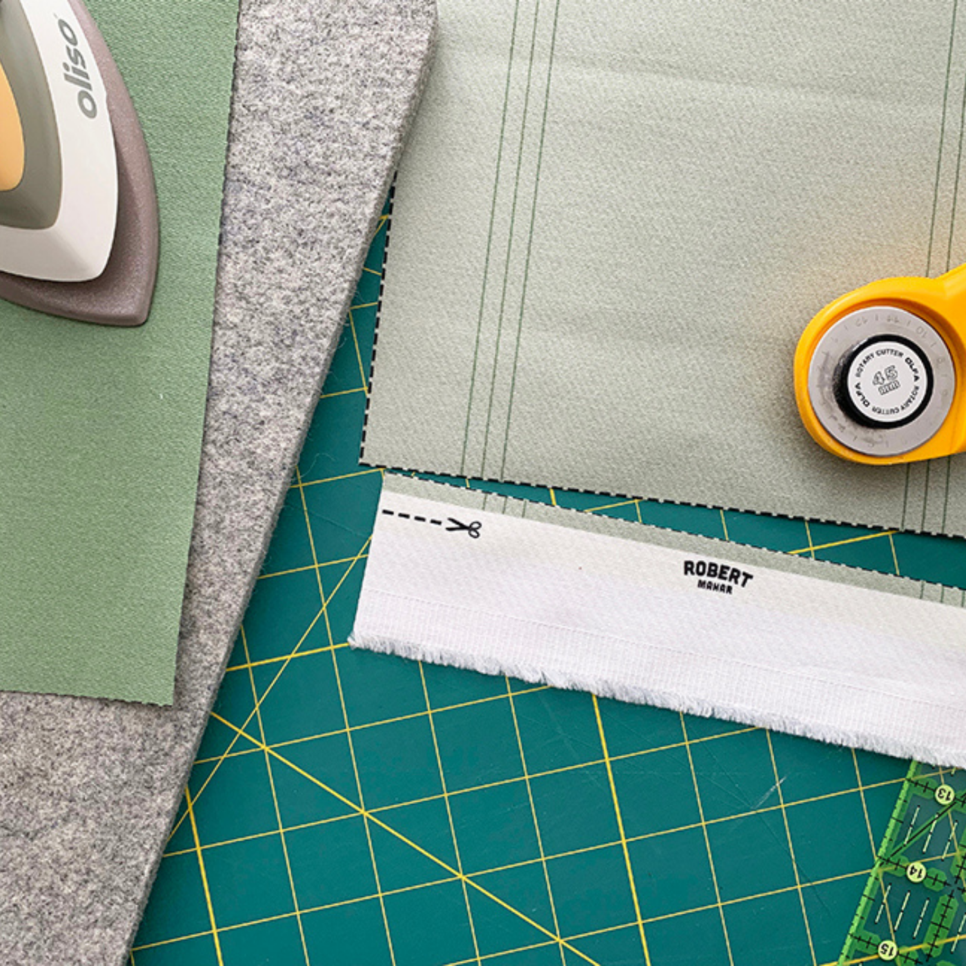A close up of some of the supplies Robert Mahar is using for his DIY holiday card display, including swatches of solid fabric in both a medium and a light green, the latter with the selvedge cut off, along with a rotary cutter, an iron and a cutting mat.