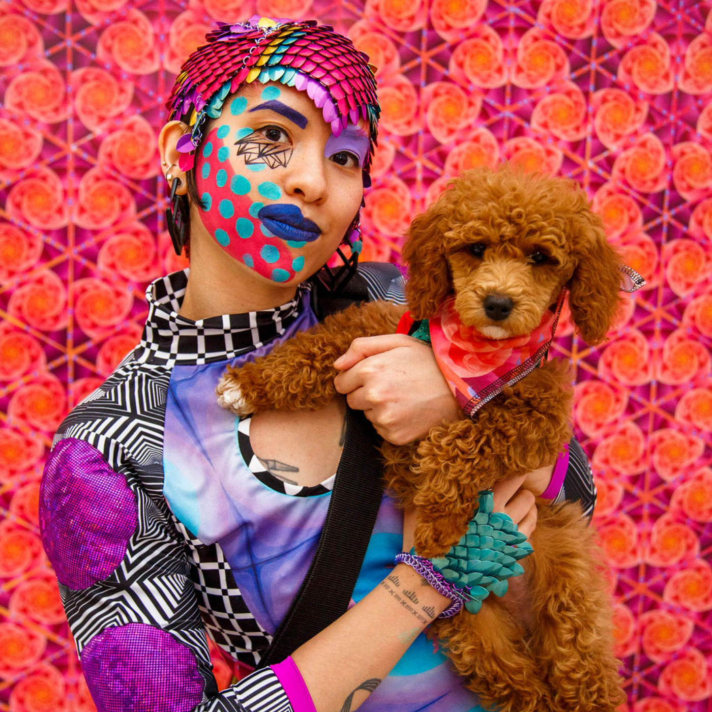 Sky Cubacub stands looking at the camera in front of a bright orange and magenta wall with swirls, holding their dog, D.O.G., who is wearing a red bandana.