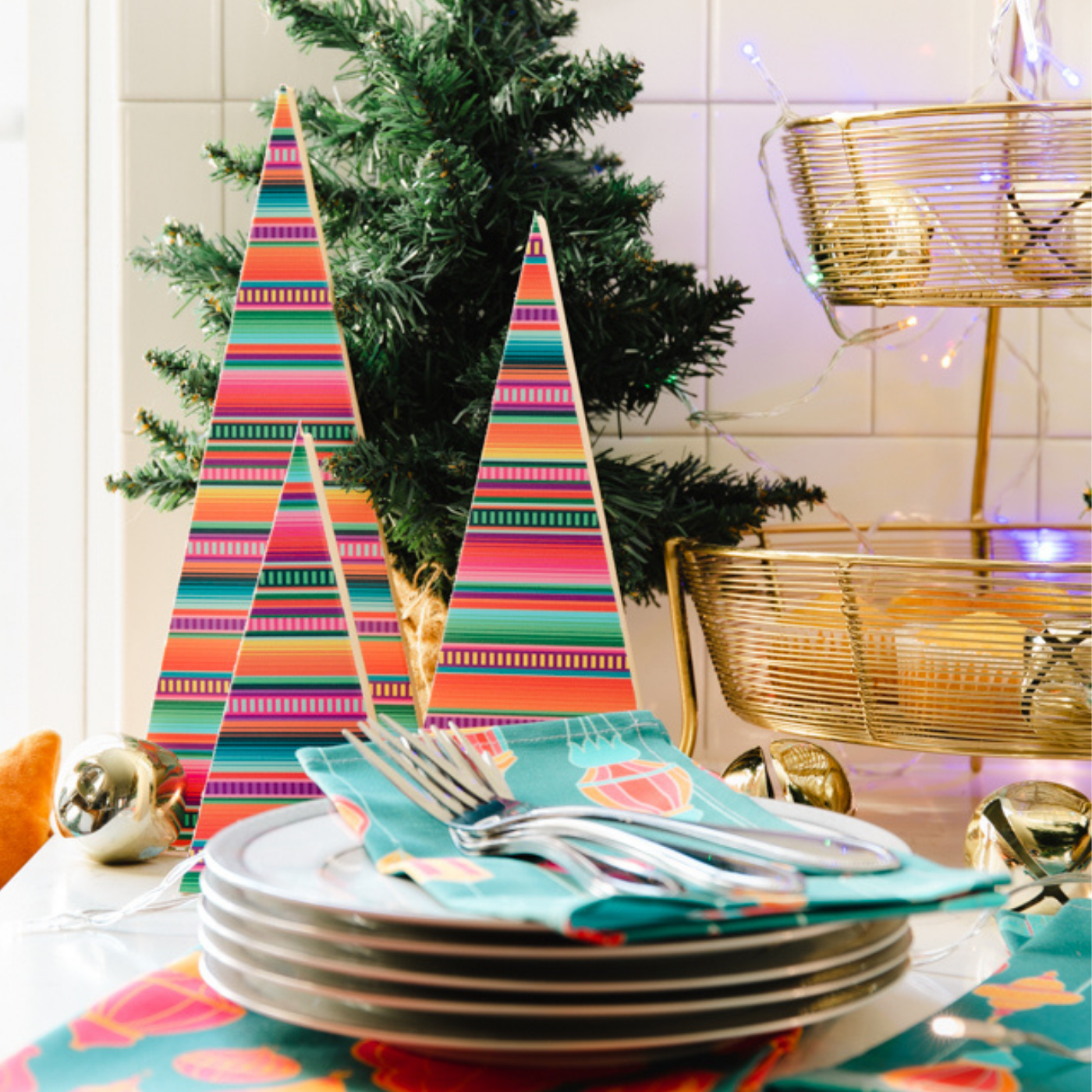 Three small Christmas trees, all comprised of triangles made out of wood, with short-width bottoms and long sides, are covered in pick-and-stick wallpaper with a brightly colored design with pink, red, blue, yellow and green horizontal stripes. The trees are part of a tablescape and placed in front a small tabletop artificial Christmas tree and behind a stack of 5 small white plates that has teal napkins with pink-and-orange-striped napkins on top of it. Several silver forks are placed on top of the napkins.